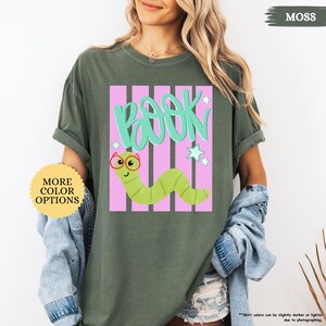 May include: A moss green short-sleeve t-shirt featuring a graphic print of a light green worm wearing red glasses. Above the worm, the word BOOK is written in teal script. The design includes pink vertical stripes and small white stars.