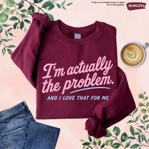 K&ouml;nnte beinhalten: Burgunderfarbenes Sweatshirt mit dem Text "I'm actually the problem. AND I LOVE THAT FOR ME" in Rosa und Hellblau. Eine Jeans und eine Tasse Kaffee sind ebenfalls im Bild.
