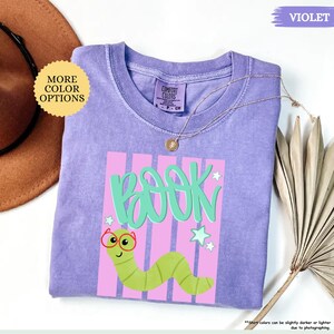 May include: A violet short-sleeved t-shirt featuring a graphic of a green worm wearing red glasses. Above the worm, the word BOOK is written in a light green script font. The design includes pink vertical stripes and small light green stars.