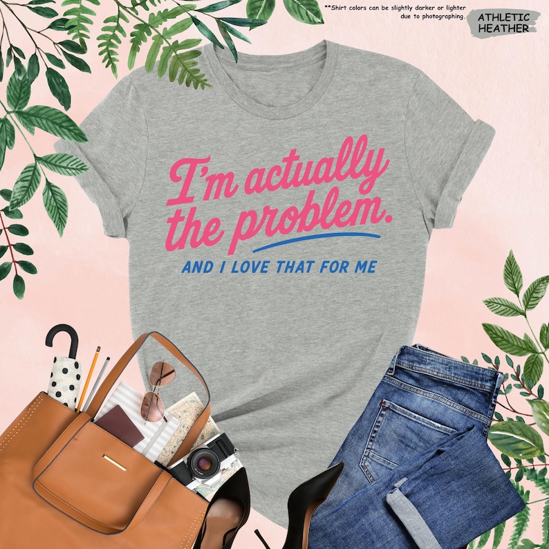 K&ouml;nnte beinhalten: Graues T-Shirt mit dem Text "I'm actually the problem. AND I LOVE THAT FOR ME" in Pink und Blau. Das Shirt wird mit einer blauen Jeans, einer braunen Handtasche und schwarzen High Heels pr&auml;sentiert.