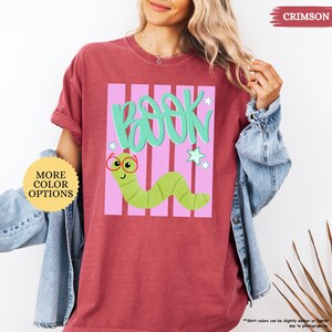 May include: A crimson t-shirt featuring a graphic of a light green worm wearing red glasses. Above the worm, the word "BOOK" is written in teal script. The design includes light pink vertical stripes and small teal stars on a white background.