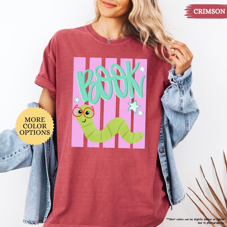 May include: A crimson t-shirt featuring a graphic of a light green worm wearing red glasses. Above the worm, the word "BOOK" is written in teal script. The design includes light pink vertical stripes and small teal stars on a white background.