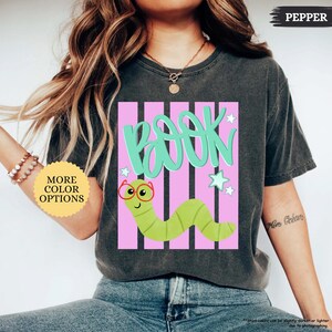 May include: A charcoal gray short-sleeve t-shirt featuring a graphic print of a green worm wearing red glasses. The design includes the word BOOK in large teal letters over vertical pink stripes and small teal stars on a dark gray background.