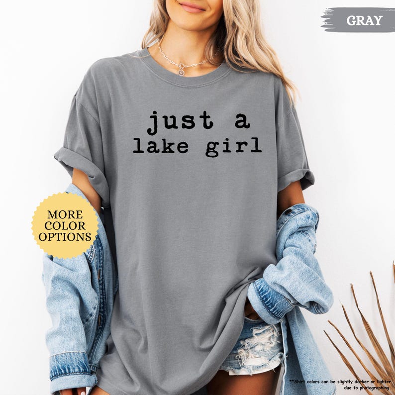 May include: Gray t-shirt with the text "just a lake girl" in black. The shirt has rolled-up sleeves and is paired with a denim jacket. A yellow sticker with "MORE COLOR OPTIONS" is visible.