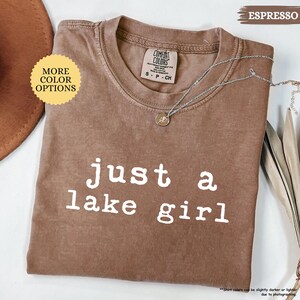 May include: Espresso-colored t-shirt with the text "just a lake girl" in white. The shirt has a crew neck and short sleeves. A silver necklace with a pendant is draped on the shirt. The shirt is labeled "Comfort Colors".