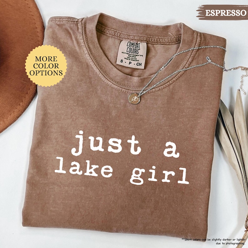 May include: Espresso-colored t-shirt with the text "just a lake girl" in white. The shirt has a crew neck and short sleeves. A silver necklace with a pendant is draped on the shirt. The shirt is labeled "Comfort Colors".