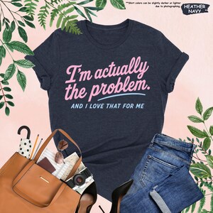 K&ouml;nnte beinhalten: Marineblaues T-Shirt mit dem Text "I'm actually the problem. AND I LOVE THAT FOR ME" in Rosa und Hellblau. Das Bild zeigt eine braune Tragetasche, eine Sonnenbrille, eine Kamera und eine blaue Jeans.