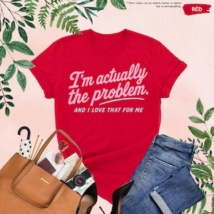 K&ouml;nnte beinhalten: Ein rotes T-Shirt mit dem Text "I'm actually the problem. AND I LOVE THAT FOR ME" in hellrosa Schrift. Das Shirt wird mit einer blauen Jeans, einer braunen Tragetasche, einer Sonnenbrille und einer Kamera kombiniert. Gr&uuml;nes Laub umgibt die Artikel.