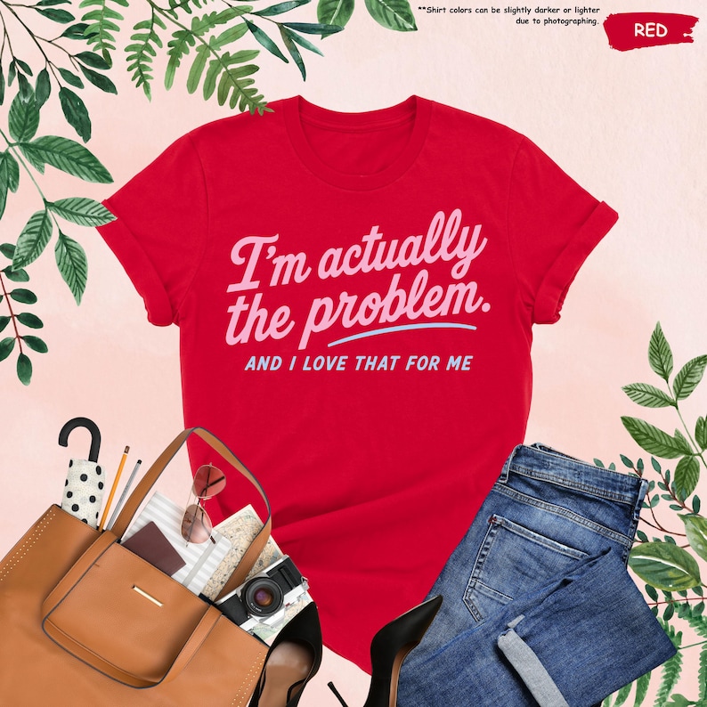 K&ouml;nnte beinhalten: Ein rotes T-Shirt mit dem Text "I'm actually the problem. AND I LOVE THAT FOR ME" in hellrosa Schrift. Das Shirt wird mit einer blauen Jeans, einer braunen Tragetasche, einer Sonnenbrille und einer Kamera kombiniert. Gr&uuml;nes Laub umgibt die Artikel.