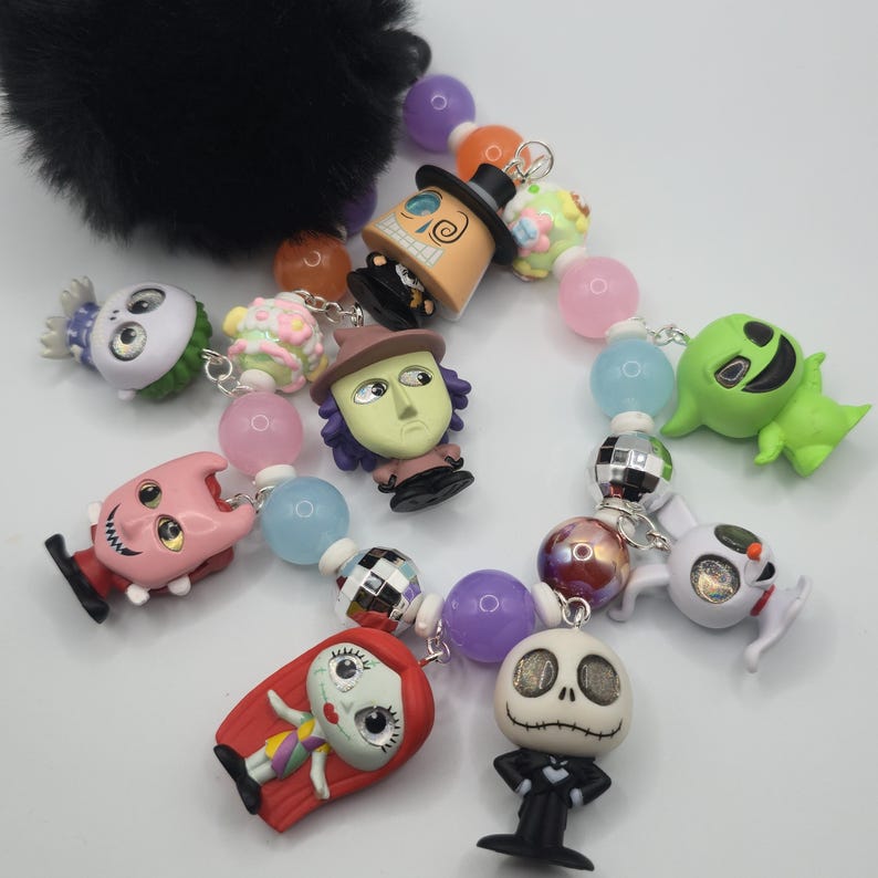 May include: A colorful beaded keychain featuring various character charms. The charms include a pink creature, a green ghost, and a Jack Skellington figure. The beads are in shades of pink, purple, and blue. A black fluffy accessory is attached to the top.