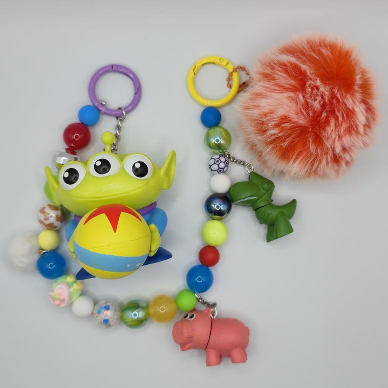 May include: A decorative keychain featuring a green three-eyed alien figure holding a yellow ball with a red star, connected to a string of multicolored beads, a small green dinosaur charm, a pink pig charm, and a large fluffy orange pom-pom.