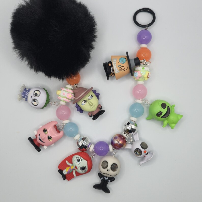 May include: A black fluffy pom-pom keychain with a beaded charm featuring characters from The Nightmare Before Christmas. The charm includes Jack Skellington, Sally, and other characters in a colorful design. The keychain is a fun accessory.