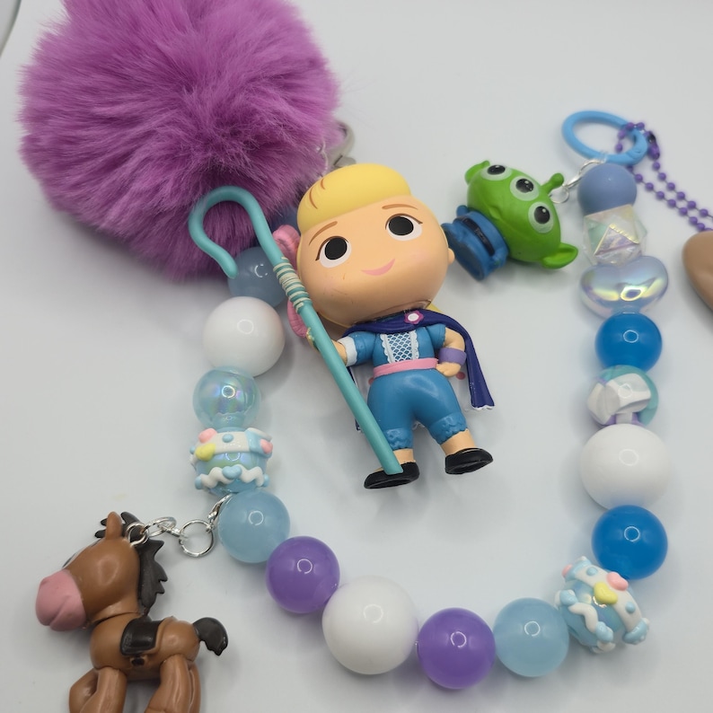 May include: A beaded keychain featuring a blonde character figurine in a blue outfit holding a light blue staff, a green three-eyed alien charm, and a brown horse charm. The accessory includes a large purple pom-pom and various blue, white, and purple beads.