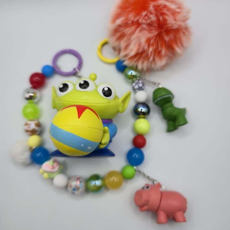 May include: A keychain featuring a green three-eyed alien figure holding a yellow ball with blue and red accents. The keychain includes a variety of colorful beads, a large orange pom-pom, and two small character charms shaped like a dinosaur and a hippo.