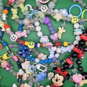 May include: A collection of colorful beaded necklaces and keychains with various charms. Charms include smiley faces, stars, rainbows, bears, hearts, and cartoon characters like Minnie Mouse and Hello Kitty. The beads are in various colors and shapes.