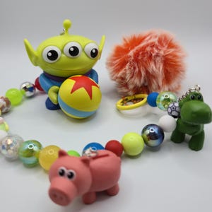 May include: A beaded keychain featuring a green three-eyed alien figure holding a yellow ball with a red star, a pink pig figure, a green dinosaur figure, and a fluffy orange pom-pom. The beads are multicolored, including translucent, metallic, and patterned spheres.