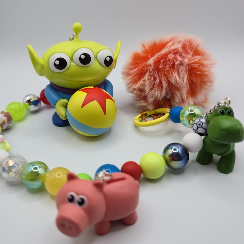 May include: A beaded keychain featuring a green three-eyed alien figure holding a yellow ball with a red star, a pink pig figure, a green dinosaur figure, and a fluffy orange pom-pom. The beads are multicolored, including translucent, metallic, and patterned spheres.