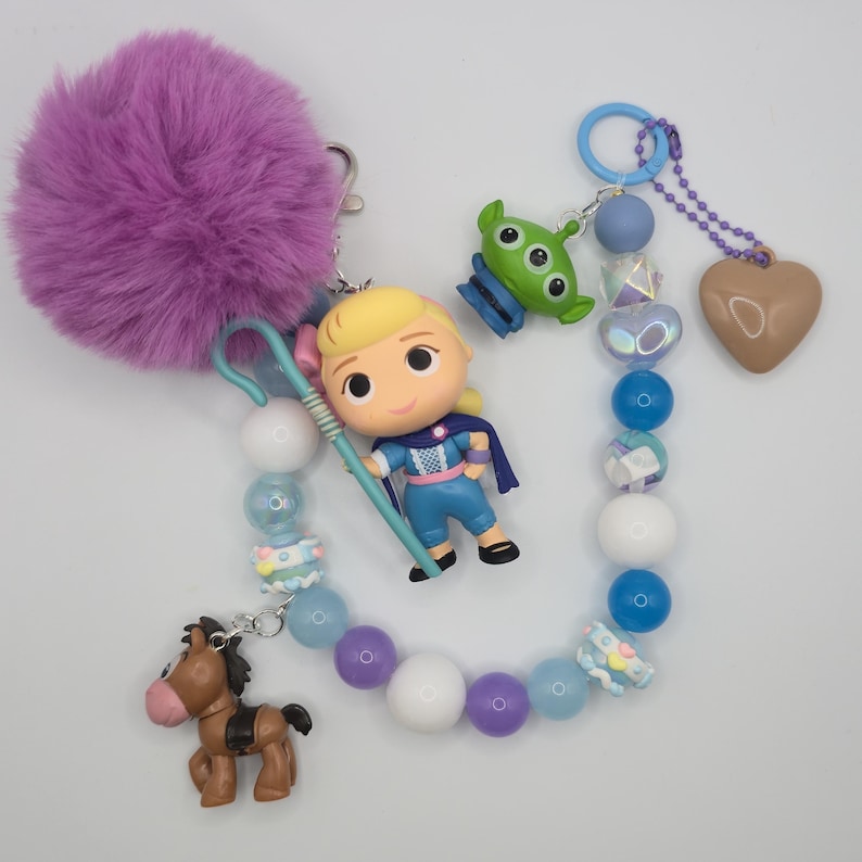 May include: A keychain featuring a purple faux fur pom-pom, a blonde character figurine with a blue shepherd's crook, a green three-eyed alien charm, a brown horse figurine, and a heart charm, all connected by a strand of blue, white, and purple beads.