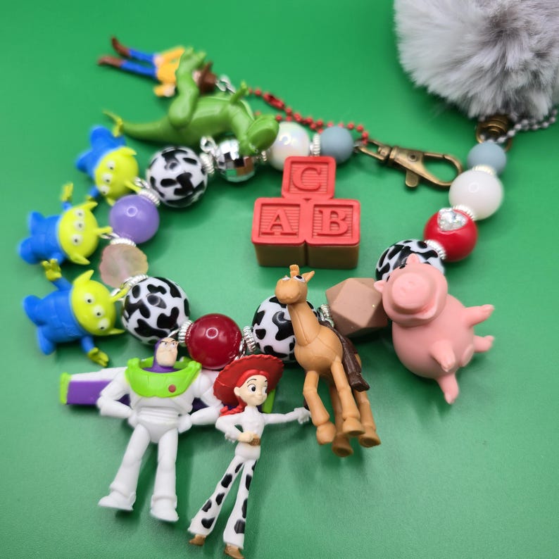 May include: A beaded keychain featuring miniature character figures including a green dinosaur, three blue aliens, a space ranger, a cowgirl, a brown horse, and a pink pig. The accessory includes red alphabet blocks with the letters C, A, and B, plus a gray pom-pom.