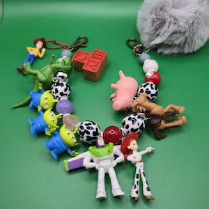 May include: A keychain accessory featuring a collection of miniature character figures including a space ranger, cowgirl, green aliens, a dinosaur, a pig, and a horse. The design incorporates black and white spotted beads, red beads, and a grey pom-pom.