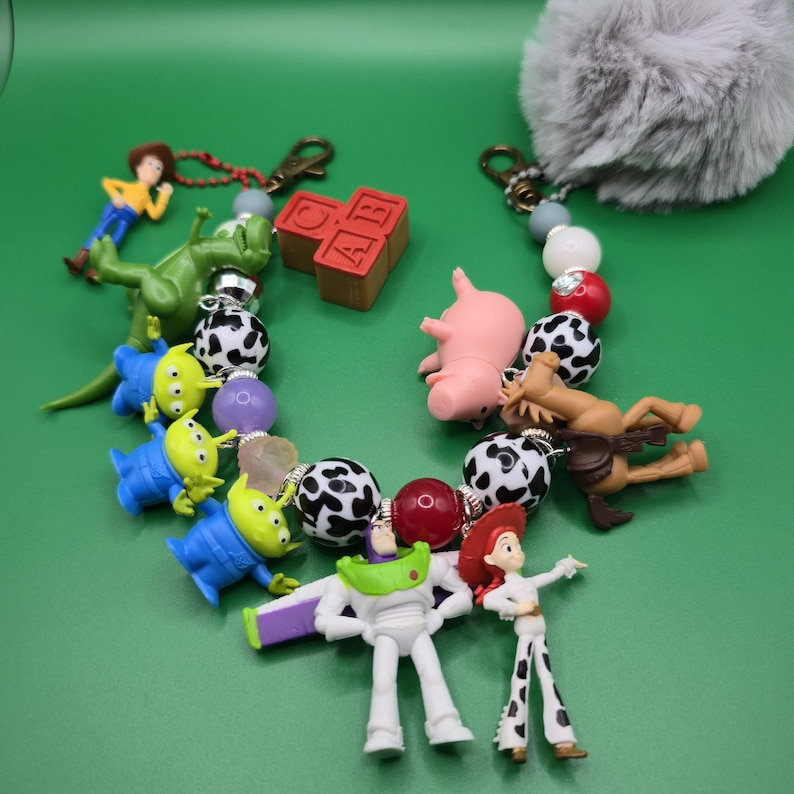 May include: A keychain accessory featuring a collection of miniature character figures including a space ranger, cowgirl, green aliens, a dinosaur, a pig, and a horse. The design incorporates black and white spotted beads, red beads, and a grey pom-pom.