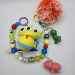 May include: A keychain featuring a green three-eyed alien figure holding a yellow ball with a red star, surrounded by a string of colorful beads, a fluffy orange pom-pom, and small green dinosaur and pink pig character charms on metal rings.