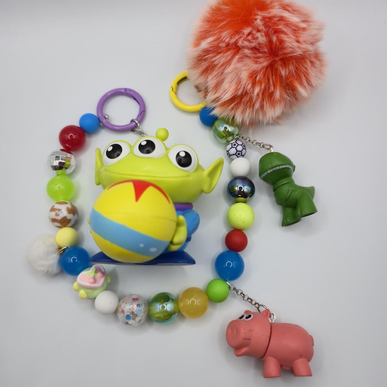 May include: A keychain featuring a green three-eyed alien figure holding a yellow ball with a red star, surrounded by a string of colorful beads, a fluffy orange pom-pom, and small green dinosaur and pink pig character charms on metal rings.