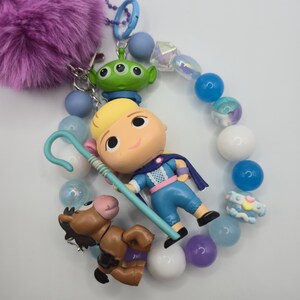 May include: A keychain featuring a purple faux fur pom-pom, a blue metal clasp, and a string of beads in shades of blue, white, and purple. The charm includes a blonde character figurine with a blue shepherd's crook, a green alien, and a brown horse.