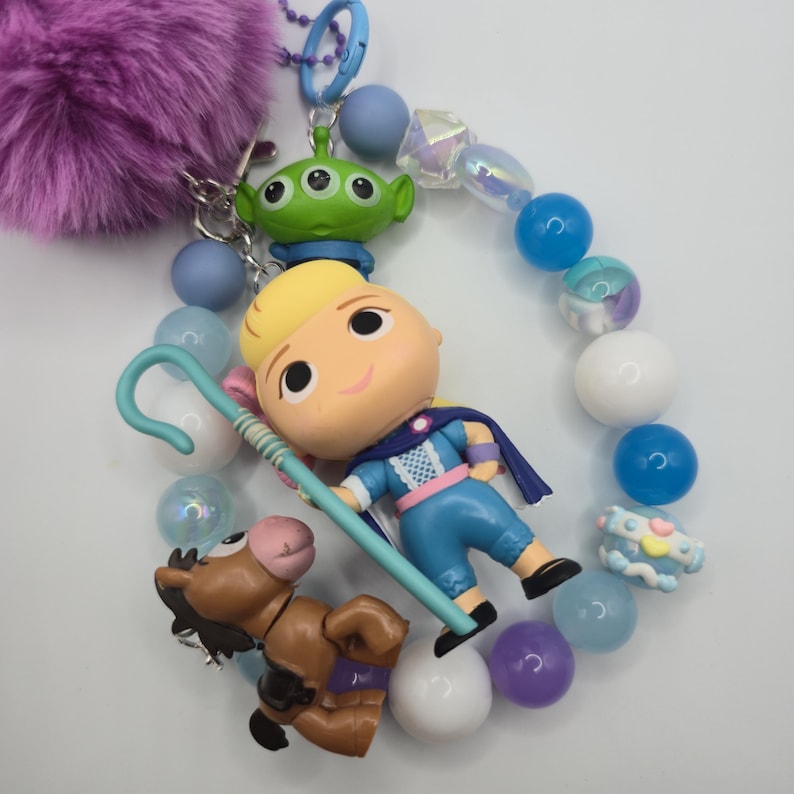 May include: A keychain featuring a purple faux fur pom-pom, a blue metal clasp, and a string of beads in shades of blue, white, and purple. The charm includes a blonde character figurine with a blue shepherd's crook, a green alien, and a brown horse.