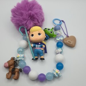 May include: A beaded keychain featuring a Bo Peep figurine with a blue shepherd's crook, a green alien character, and a brown horse figure. The strand includes purple, blue, and white beads, a purple pom-pom, and a brown heart-shaped charm.