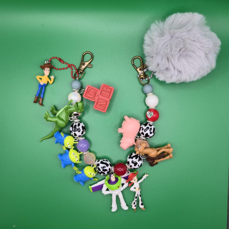 May include: A beaded keychain featuring miniature character figures including a cowboy, a space ranger, a cowgirl, a green dinosaur, a pig, and three green aliens. The keychain includes black and white cow-print beads, a red block with letters CAB, and a large grey faux fur pom-pom attached to a metal lobster clasp.
