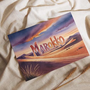 Könnte beinhalten: Aquarelldruck mit einer Wüstenlandschaft und dem Wort "Marokko" in Orange und Gelb. Das Kunstwerk zeigt einen Sonnenuntergang über Sanddünen mit violetten und orangen Farbtönen. Der Druck befindet sich auf einem weißen Hintergrund.