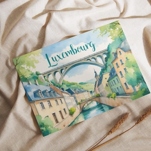 May include: A watercolor postcard featuring a scenic view of Luxembourg. The image showcases a bridge, buildings, and a river, with the word "Luxembourg" in teal script. The postcard is set against a beige fabric background.