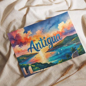 Puede incluir: Una impresión en acuarela de Antigua, que representa una puesta de sol sobre el mar con veleros y colinas verdes. La palabra "Antigua" está escrita en azul sobre la imagen.