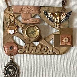 May include: A gold-toned necklace with a square pendant featuring a compass, a small eagle, and the word "Arles". The pendant is made of various metal pieces and has a vintage look. A small portrait charm hangs from the bottom of the pendant.