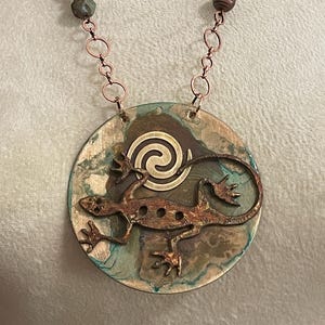 May include: A handcrafted necklace featuring a circular pendant with a copper lizard design and a spiral accent. The pendant has a mottled blue and brown background. The necklace includes copper chain links and beaded accents.