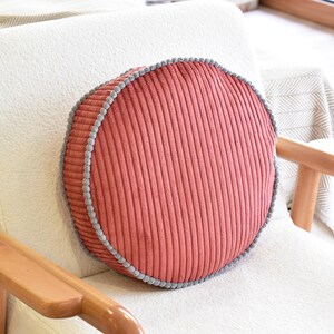 May include: A round, terracotta-colored corduroy pillow featuring a textured, ribbed fabric surface. The circular cushion is finished with a decorative grey pom-pom trim that runs along the entire outer edge, providing a distinct contrast.