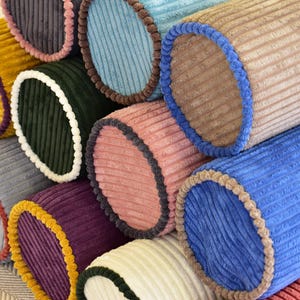 May include: A close-up of a stack of colorful, corduroy fabric rolls. The rolls are in various colors, including yellow, green, pink, blue, and purple. Each roll has a contrasting border, creating a visually appealing display.