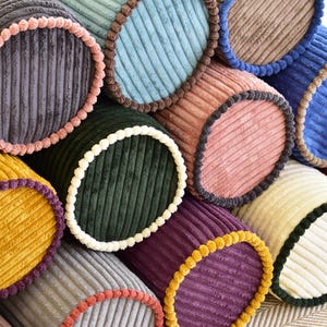 Op de afbeelding: Een verzameling kleurrijke bolsterkussens van corduroy in tinten geel, grijs, groen, blauw, roze en paars. Elk kussen heeft een contrasterende rand, wat een aantrekkelijk spel van texturen en kleuren creëert.
