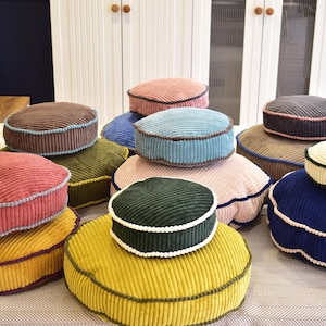 May include: A collection of colorful, round corduroy cushions in various shades, including red, blue, green, and yellow. Each cushion has a contrasting border. The cushions are stacked, creating a visually appealing display.