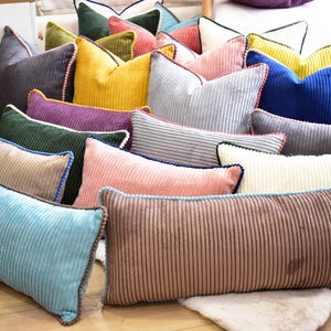 May include: A collection of decorative throw pillows in various colors and patterns, including blue, green, yellow, pink, and brown. The pillows are made of a corduroy fabric with a ribbed texture and have a decorative trim around the edges.