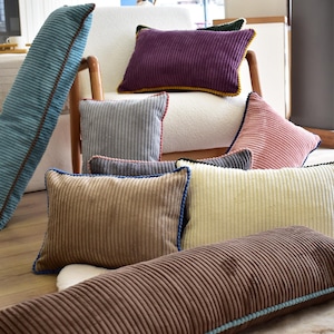 Puede incluir: Una colección de cojines de pana en varias formas y colores, incluyendo azul, morado, gris, rosa, marrón y crema. Los cojines tienen ribetes contrastantes y están colocados sobre un suelo de madera y una silla blanca.