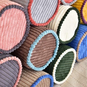 Pode incluir: Uma coleção de almofadas cilíndricas de veludo cotelê coloridas. As almofadas têm uma textura canelada e acabamento contrastante. As cores incluem rosa, cinza, marrom, azul, verde e amarelo. A borda tem uma cor diferente da almofada.