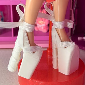 May include: White platform heels with ribbon ankle ties. The heels are on a red, glittery base. The shoes have a textured appearance and are designed for a doll.