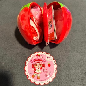May include: A red strawberry-shaped container, open to reveal contents, with a matching tag. The tag features a cartoon girl with red hair and a pink cat, with the text "Strawberry Shortcake". The container is red with green accents.