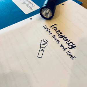 May include: A black flashlight rests on a white sheet of paper with the words "Emergency Lighting, Power, and Heat" in cursive. A blue binder labeled "Emergency Prep Binder" is in the background.