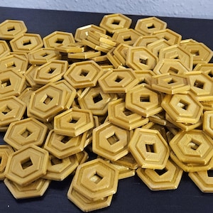 May include: A collection of gold-coloured, hexagonal game tokens. Each token features a square hole in the centre and a raised border. The tokens are stacked on a dark surface.
