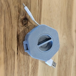 May include: A gray retractable USB cable with a white connector. The cable is wound around a gray plastic spool with a textured surface.