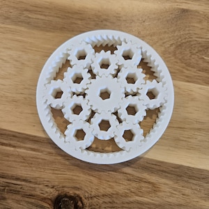 Può includere: Un set di ingranaggi in plastica bianca con più ingranaggi interbloccati di diverse dimensioni. Gli ingranaggi sono disposti in un motivo circolare.