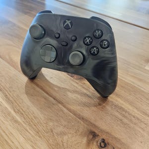 May include: A black and gray Xbox controller with a marbled design. The controller has a black Xbox logo and black buttons.